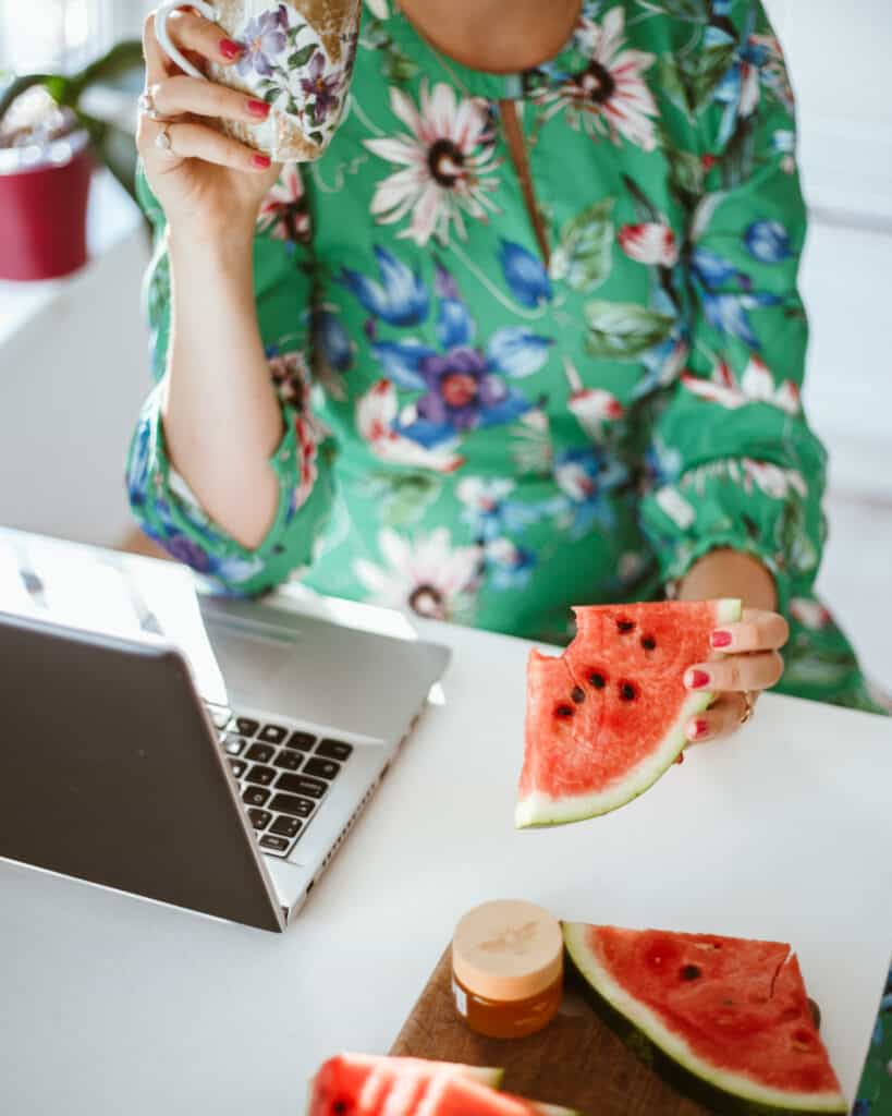 woman in dress sitting at laptop eating watermelon and drinking tea